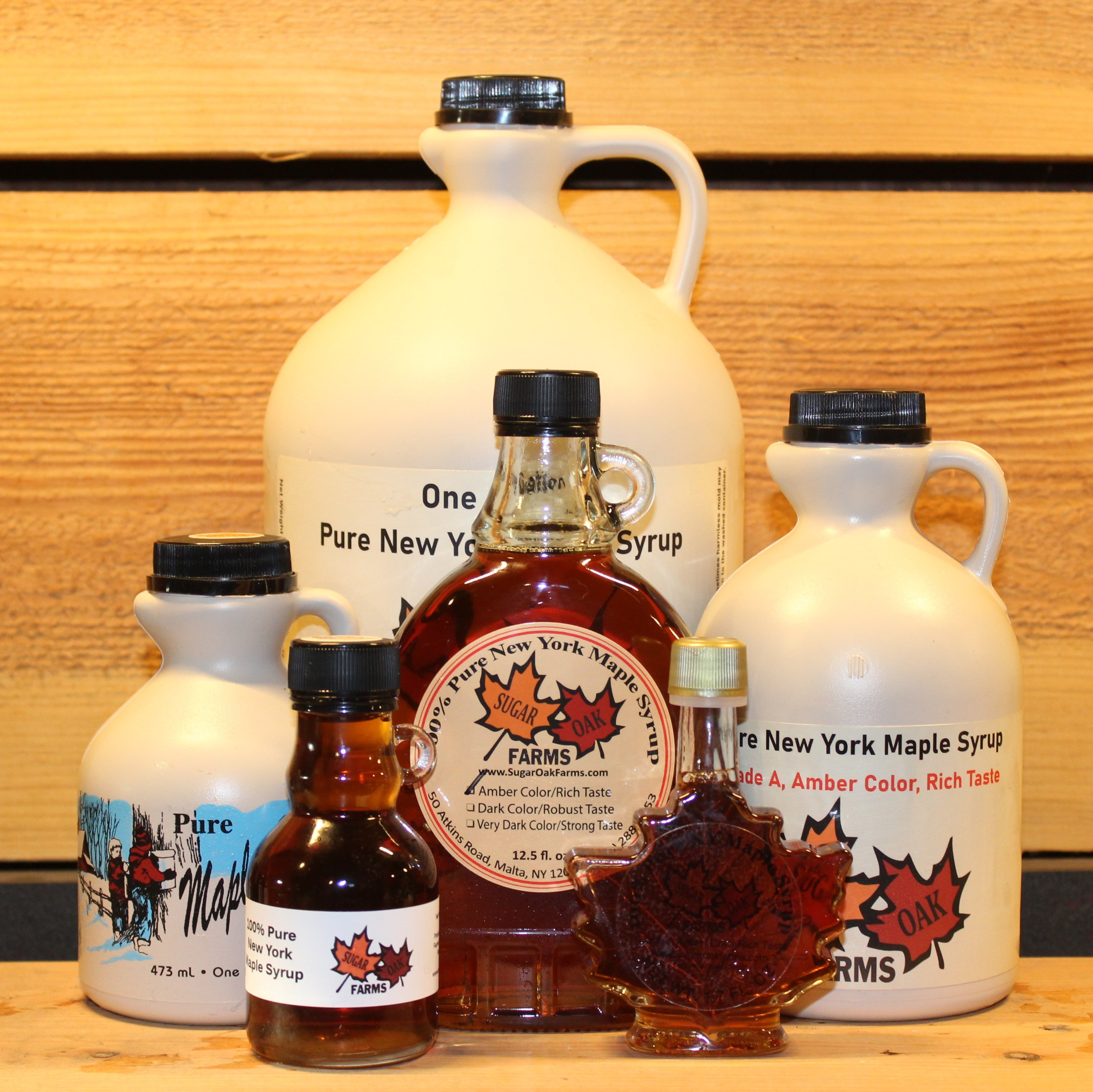 Collection of six different sizes of amber maple syrup in three rows. One gallon plastic jug of amber syrup in the back with a row of three smaller containers of syrup in front of the gallon jug and the front row has two of the smallest syrup sizes. The middle row from left to right has a plastic pint of syrup, a 12.5 fl. oz. glass bottle of syrup, and a plastic quart of syrup. The front row, from left to right, has a 8 fl. oz. glass jug of syrup and a 8.45 fl. oz. glass leaf bottle of syrup.