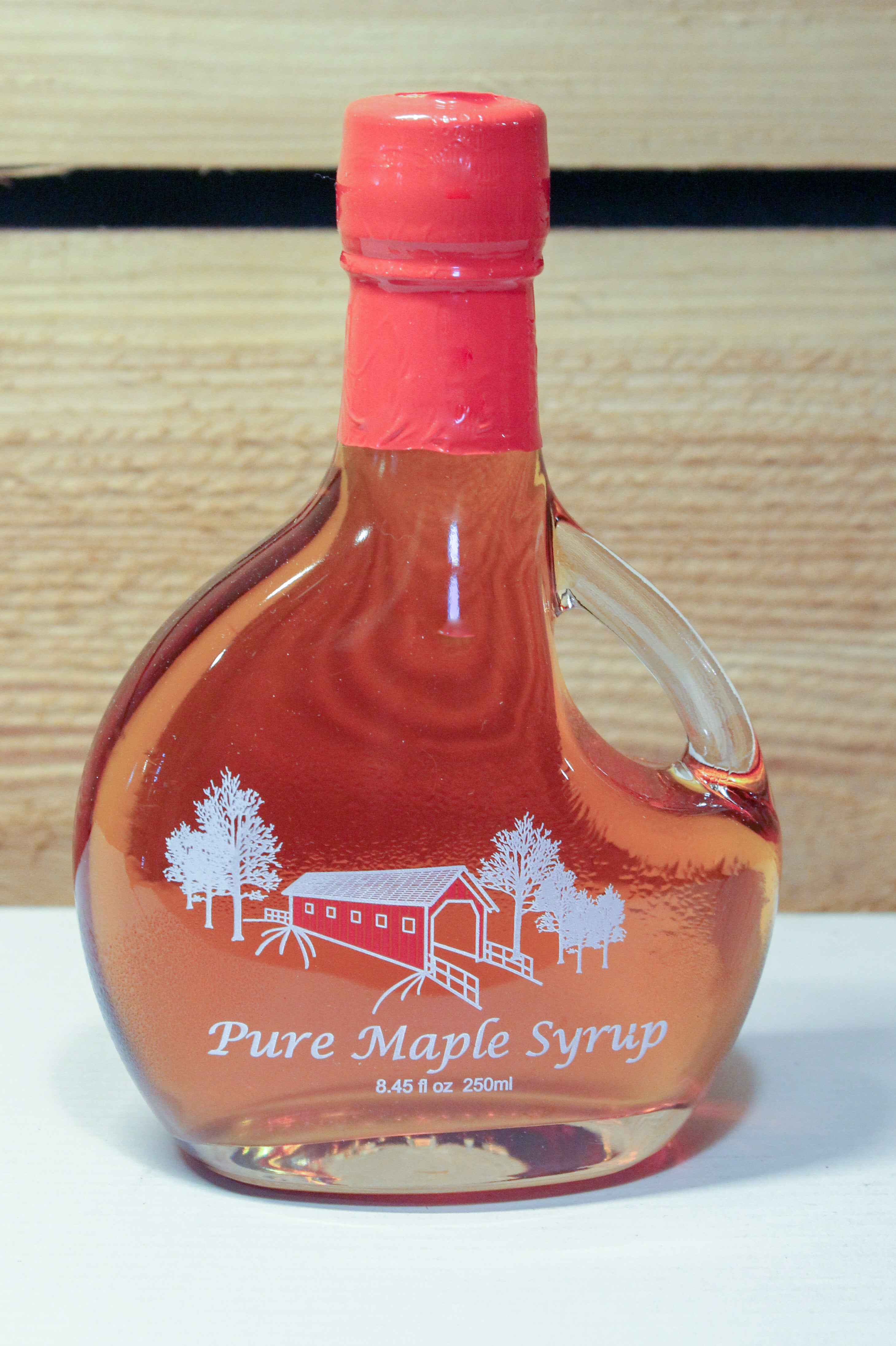 A 8.45 fl. oz. glass jug of amber syrup with a red covered bridge between white trees printed on the front.
